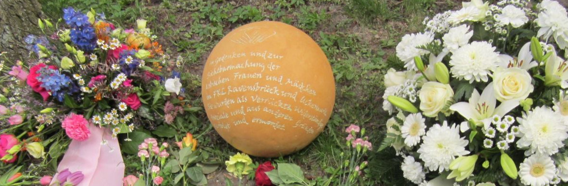 Auf dem Foto ist die Gedenkkugel für die verfolgten und ermordeten lesbischen Frauen und Mädchen im ehemaligen Frauenkonzentrationslager Ravensbrück und Uckermark zu sehen, eingerahmt von zwei Blumengestecken.